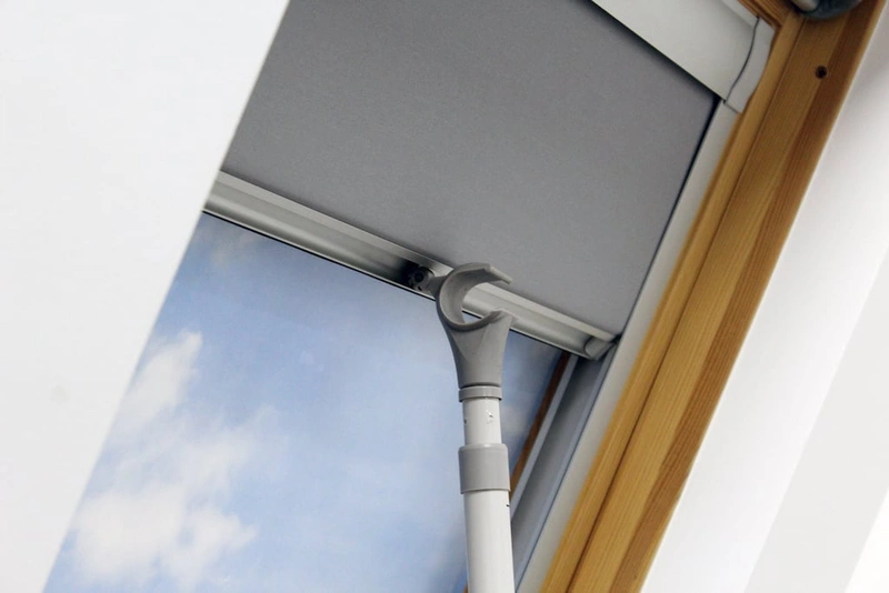 Close-up view of a skylight blind operated with a manual pole, showcasing its easy-to-use design. The sleek grey blind is installed in a natural wood-framed window, providing a practical solution for hard-to-reach windows while offering privacy and light control."