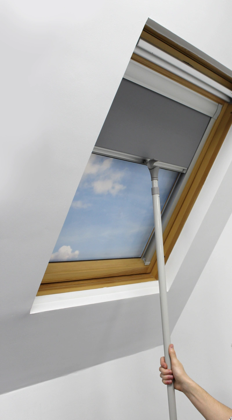 User operating a skylight blind with a manual hook pole, demonstrating ease of use for hard-to-reach windows. The sleek grey blind is set within a natural wood-framed window, combining practicality with modern aesthetics for effective light control and privacy