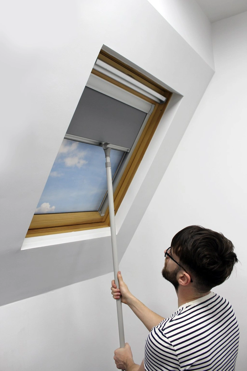 User operating a skylight blind with a manual hook pole, demonstrating ease of use for hard-to-reach windows. The sleek grey blind is set within a natural wood-framed window, combining practicality with modern aesthetics for effective light control and privacy